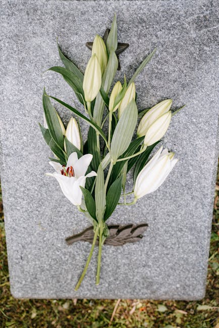 A floral arrangement of white lilies and creamy white roses, with long green leaves, arranged and placed against a grey stone surface. The lilies are in various stages of bloom, showcasing their trump