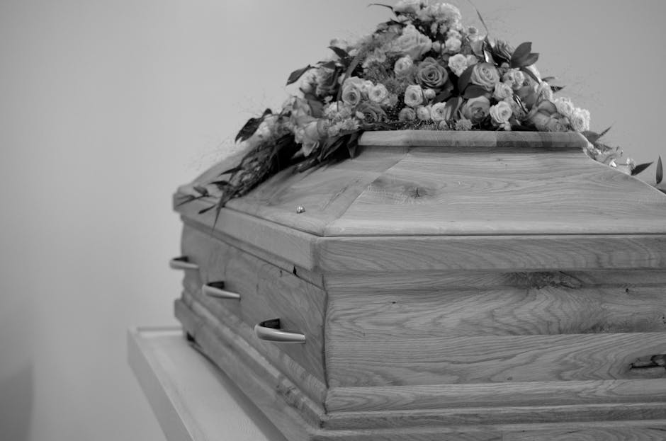 A wooden coffin with a smooth, polished surface featuring natural wood grain patterns rests on a white platform against a plain background. On top of the coffin, there is a floral arrangement consisti