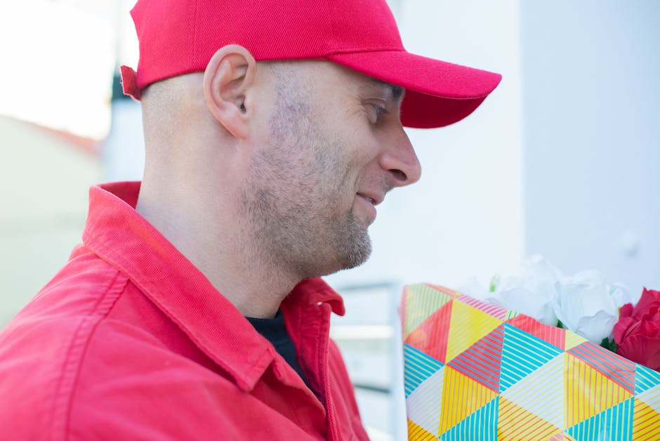 A man wearing a red cap and a matching red jacket is holding a colorful, geometric-patterned gift bag filled with fresh white and red flowers, likely roses or peonies. The arrangement features blooms 