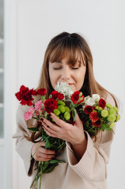 A woman with light brown hair and a gentle smile gently holds a vibrant bouquet of fresh flowers, including red carnations, pink roses, white carnations, and green chrysanthemums. The flowers are arra