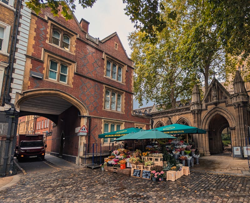 A street-side flower stall situated beneath a green canopy with the logo 'flowerdelivery.co.uk,' displaying a vibrant array of fresh flowers. The bouquet arrangements include roses, chrysanthemums, li