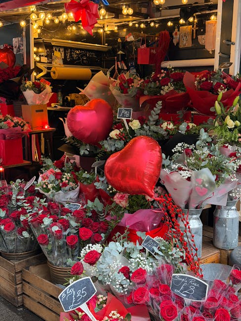 A vibrant display of floral arrangements featuring red roses, white flowers, and greenery, arranged in bouquets wrapped in pink and white paper. Among the flowers are large red heart-shaped balloons a