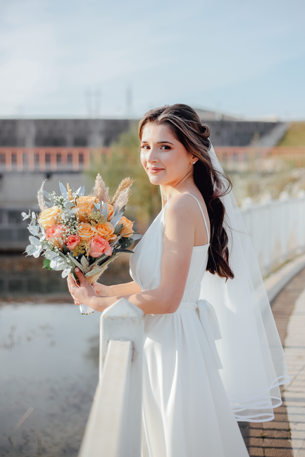 A young woman with long dark hair, dressed in a white wedding gown, is standing outdoors on a wooden pathway near Maidenhead Bridge, holding a vibrant floral bouquet. The arrangement includes peach an